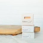 Three Eden's Gift boxed haircare products stacked on a wooden board against a white brick wall background. The top box is a blush pink color, while the two lower boxes are white with gray text reading 'BALANCED'.