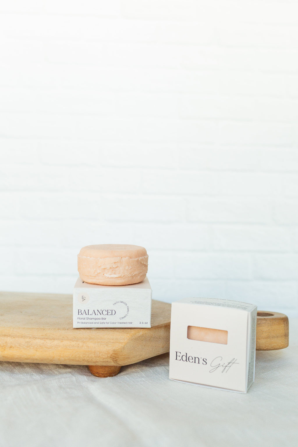 A small round peach-colored cosmetic or soap bar sits on top of a white box labeled BALANCED, next to another white box labeled Eden's Gift, arranged on a light wood board against a white brick wall background.