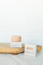 A small round peach-colored cosmetic or soap bar sits on top of a white box labeled BALANCED, next to another white box labeled Eden's Gift, arranged on a light wood board against a white brick wall background.
