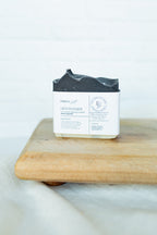 A handmade soap bar with a dark charcoal gray top and a white paper label, resting on a wooden cutting board against a white brick wall background.
