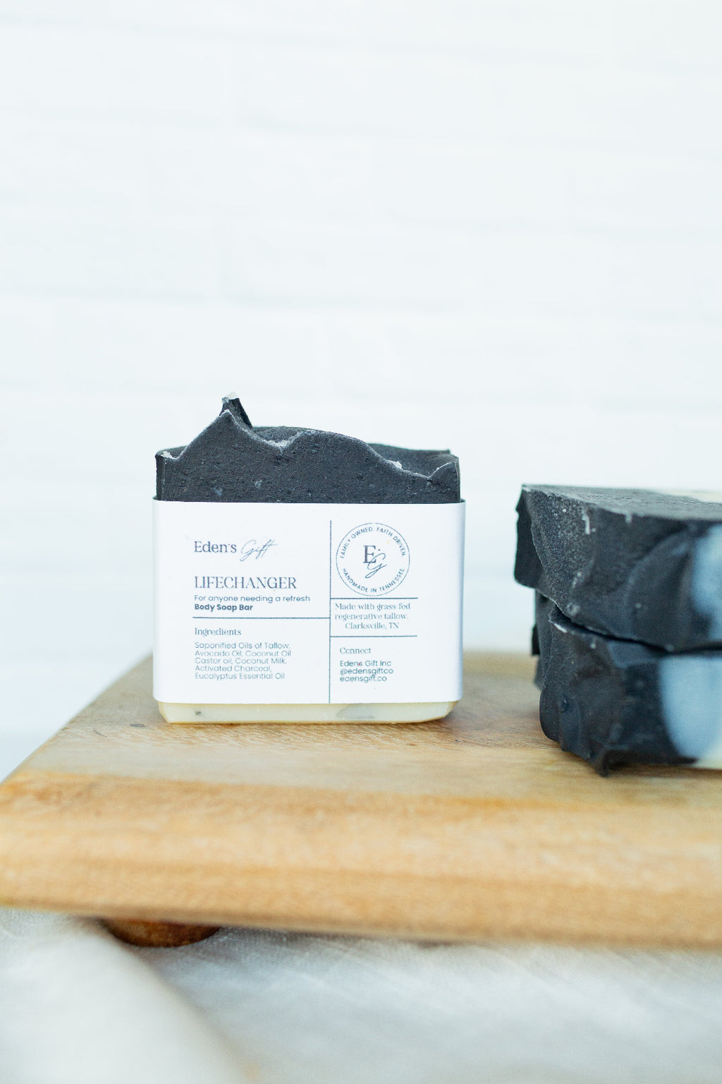 A bar of soap labeled 'Loochanger' by Edem's, wrapped in a white paper sleeve with a dark gray top, displayed on a wooden board next to another wrapped bar of soap against a white brick wall background.
