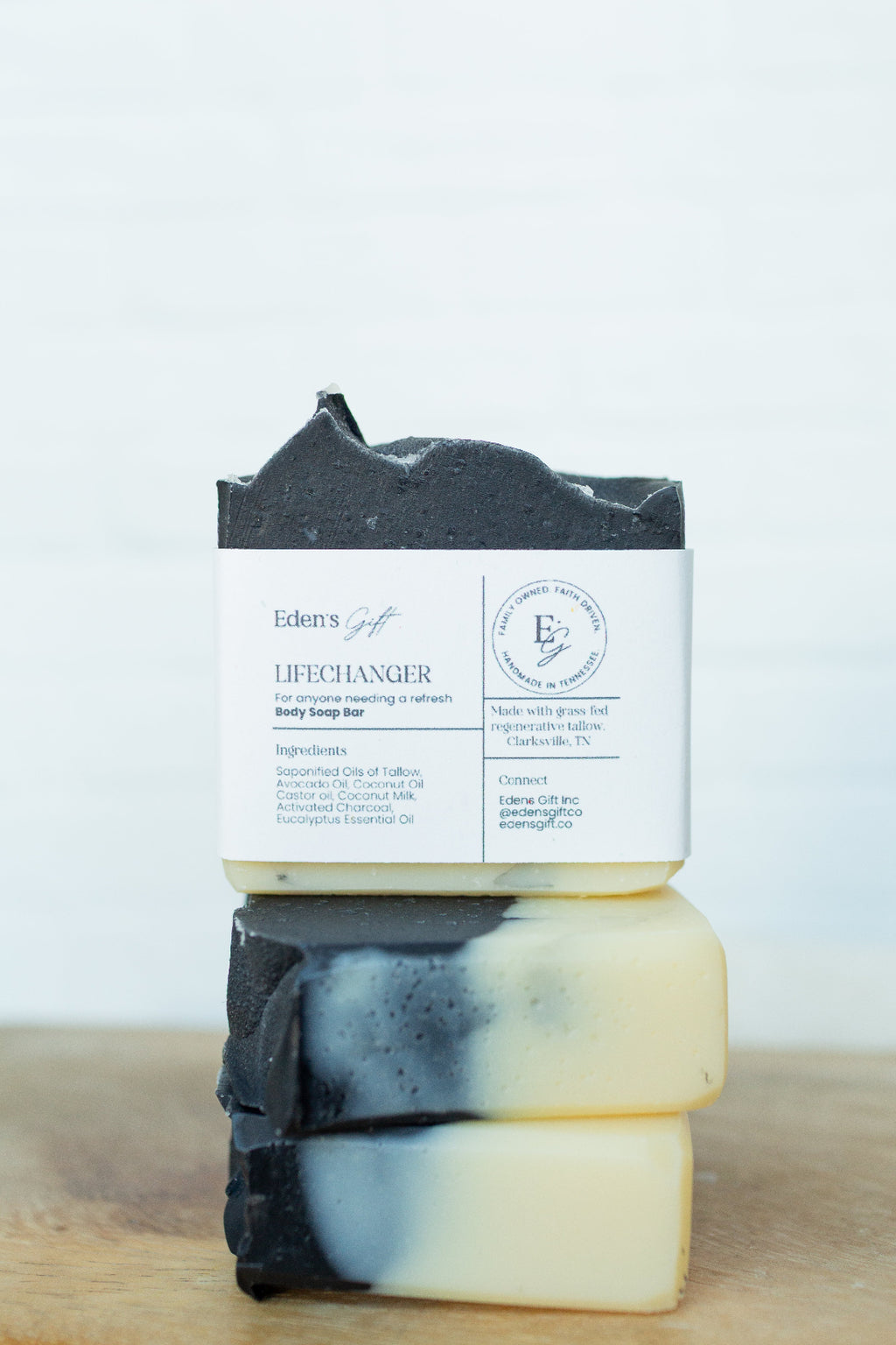 Two stacked bars of handmade soap on a wooden surface against a white background. The top bar is dark charcoal gray with a white paper label reading 'Edens of Life LIFELANGER'.