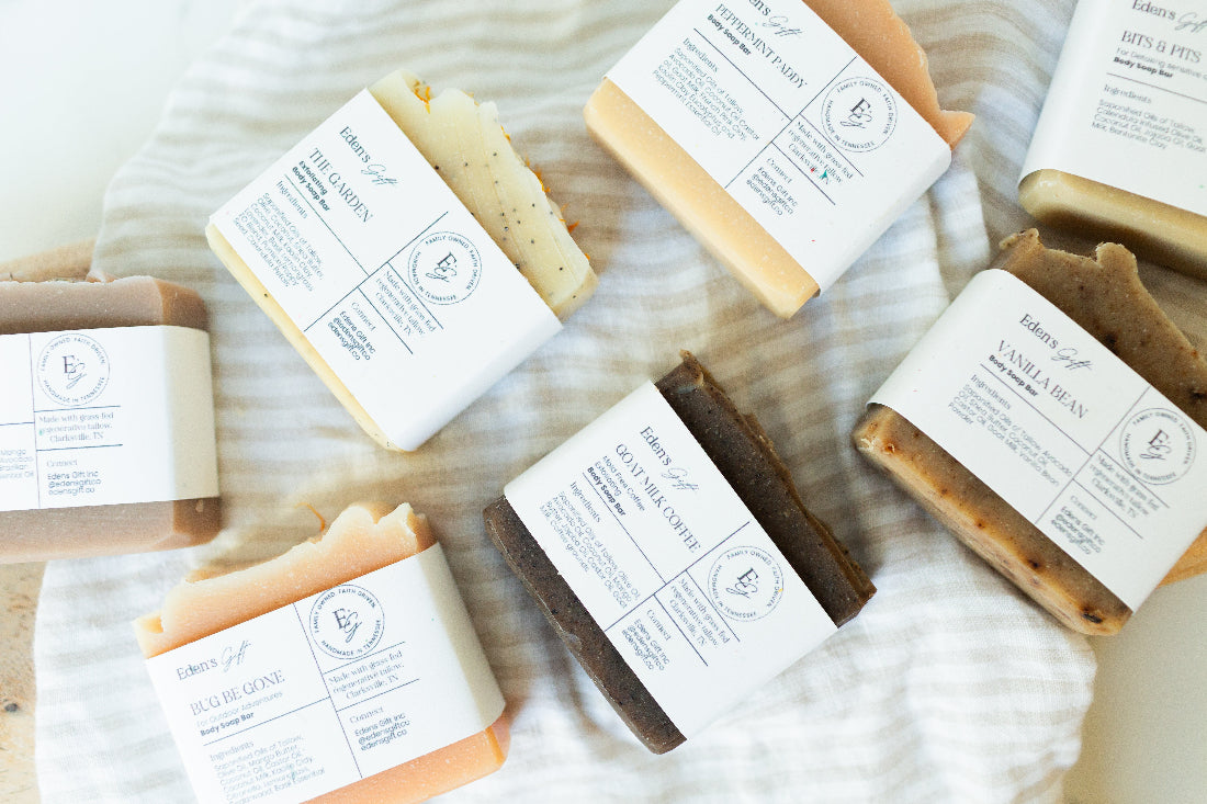 Several artisanal bars of soap wrapped in white paper labels, arranged on a white striped fabric background. The soaps come in various colors including beige, brown, orange, and cream.