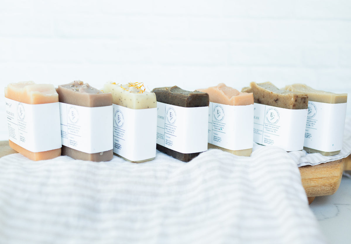 A row of artisanal handmade soaps in various earthy tones, each wrapped in a white paper band with a minimalist logo, arranged on a wooden board over a white striped towel.