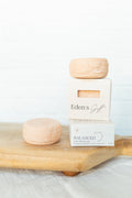 Two pale peach-colored soap bars displayed on a light wood board, with one wrapped in white paper and labeled 'Eden's Gift' and the other labeled 'BALANCED'.