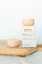 Two pale peach-colored soap bars displayed on a light wood board, with one wrapped in white paper and labeled 'Eden's Gift' and the other labeled 'BALANCED'.