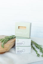 Two white and pale green boxes of Eden's Gift Growth Rosemary Enriched Handmade Soap stacked on a white cloth with rosemary sprigs and a wooden board.