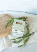 Three small white gift boxes stacked together on a light fabric surface, with rosemary sprigs and a wooden board around them. The top box is pale green and printed with the words 'Eden's Gift'.