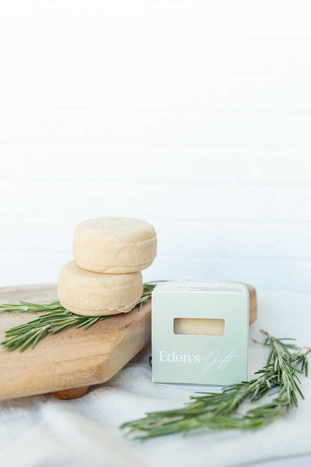 Two round pale beige soap bars stacked on a wooden board beside a mint green box labeled Eden's Gift, with rosemary sprigs on a white surface against a white wood plank background.