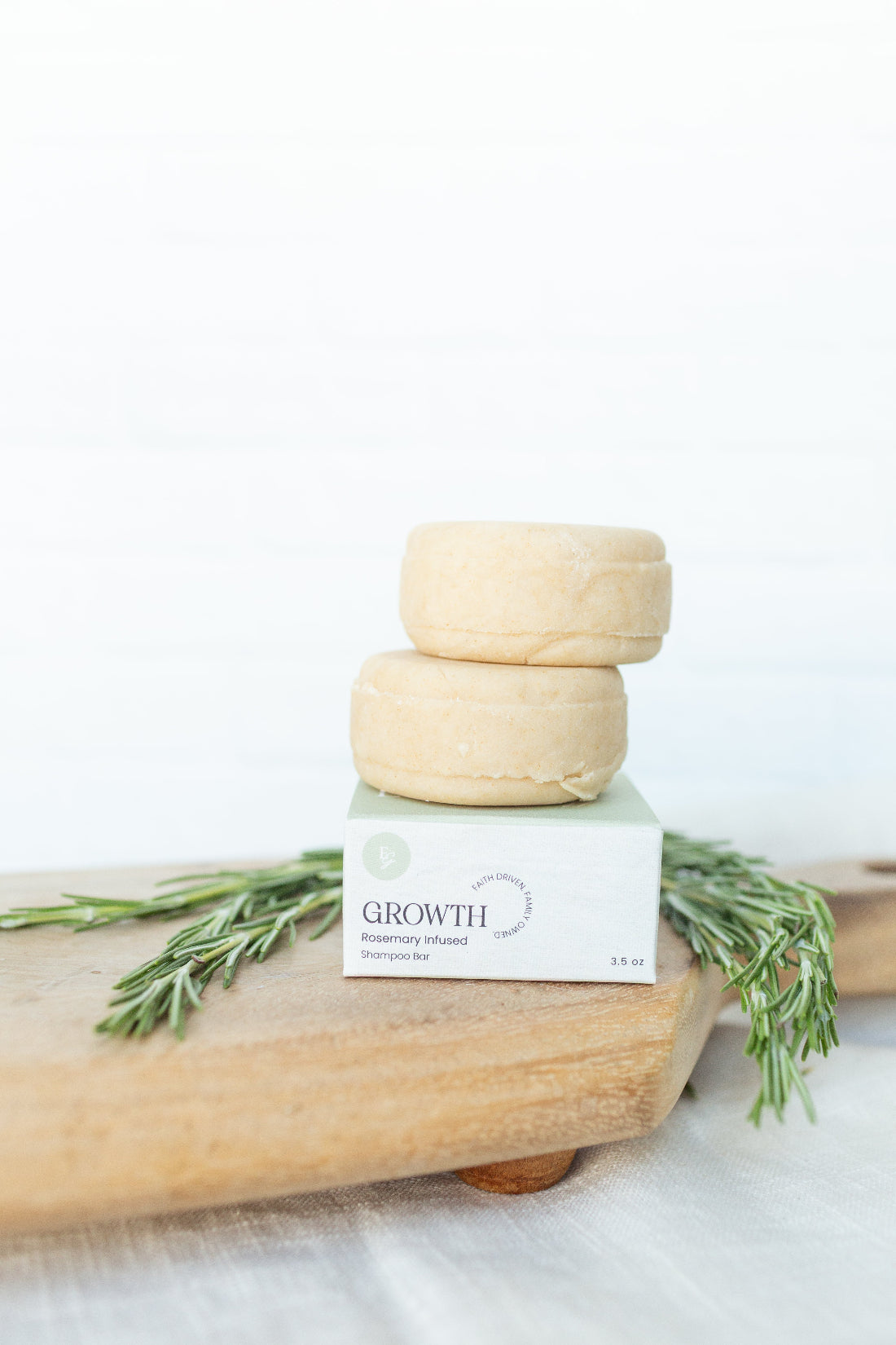 Two round beige shampoo bars stacked on top of a white box labeled GROWTH, placed on a wooden board with rosemary sprigs against a bright white background.