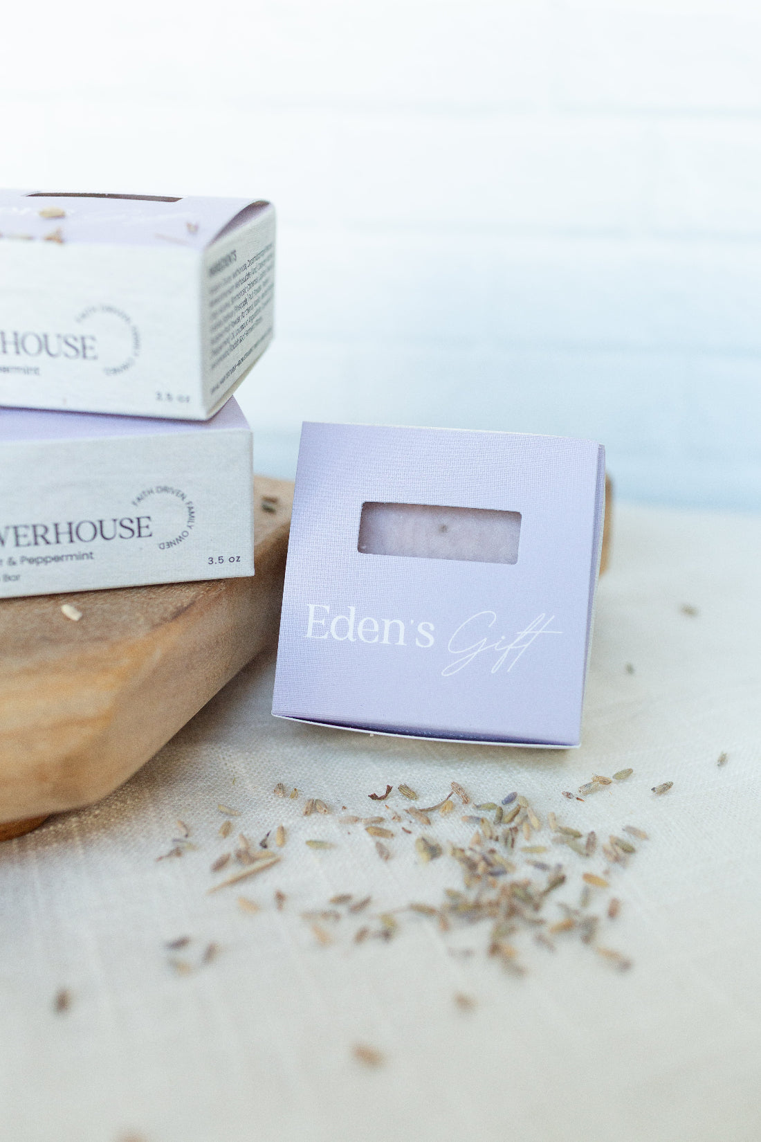 A lavender gift box with a clear window displaying a lavender soap bar, alongside two larger white and lavender boxes from Eden's Gift and Swerhouse on a wooden board with scattered dried lavender.