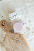 A round pale pink soap bar in a clear square container sits on a white box labeled POWERHOUSE, styled on a cream fabric surface with a wooden cutting board and dried botanicals nearby.