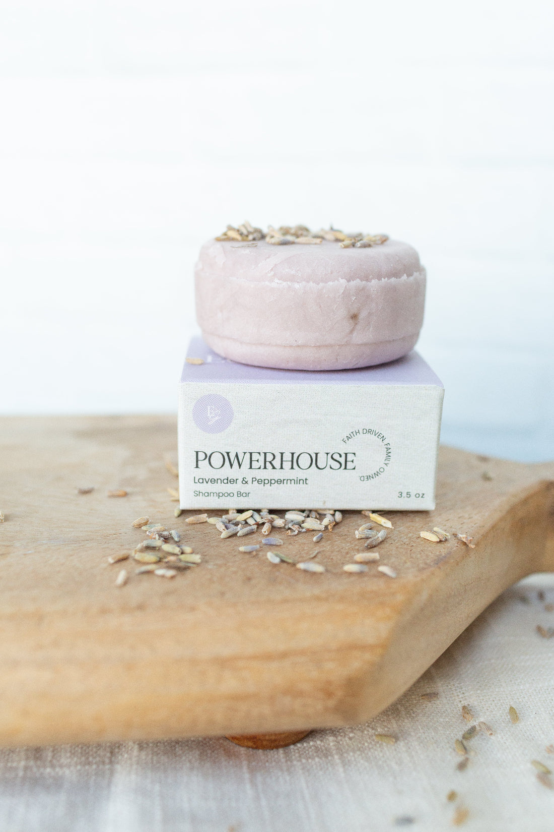 A lavender and peppermint shampoo bar by POWERHOUSE sits on top of its white and lavender packaging box, styled on a wooden cutting board with scattered dried lavender buds.