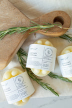 Three round bars with white labels reading 'Elder's Gift' and 'ROSEMARY' are arranged on a light fabric surface with rosemary sprigs and a wooden cutting board.