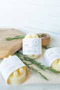 Three pale yellow wax melts in paper sleeves labeled 'HERBARY' and 'Rosemary', arranged on a light surface with rosemary sprigs and a wooden cutting board.