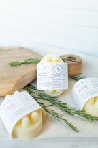 Three pale yellow wax melts in paper sleeves labeled 'HERBARY' and 'Rosemary', arranged on a light surface with rosemary sprigs and a wooden cutting board.