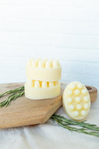 Three pale yellow massage scalpers with silicone nubs arranged on a wooden cutting board with rosemary sprigs, against a white brick wall background.