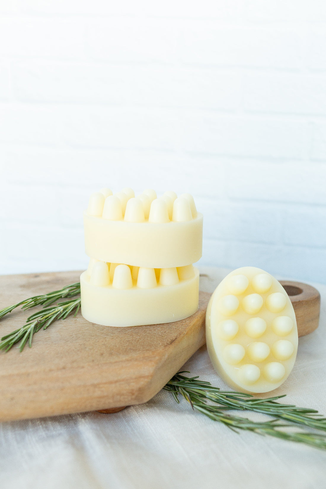 Three pale yellow massage scalpers with silicone nubs arranged on a wooden cutting board with rosemary sprigs, against a white brick wall background.