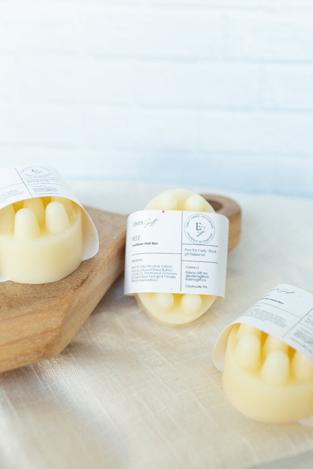 Three pale yellow wax melts in crinkle paper sleeves with white labels, arranged on a light fabric surface with a wooden board in the background.