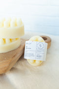 Cream-colored natural hair melt scalpels stacked on a wooden board next to a wrapped product label on a light fabric surface.